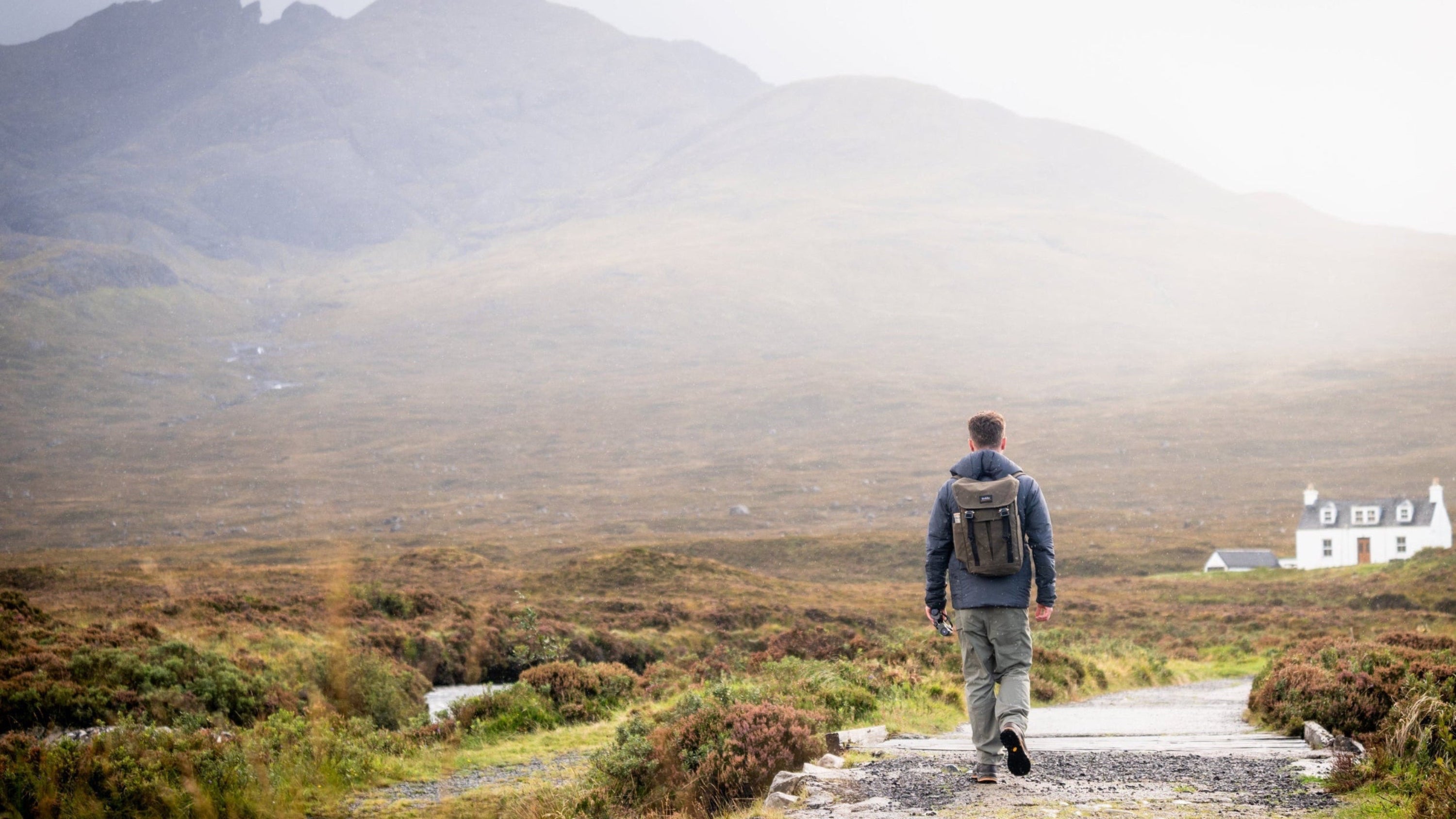 Person standing on a path looking at mountains with a house in the distance.